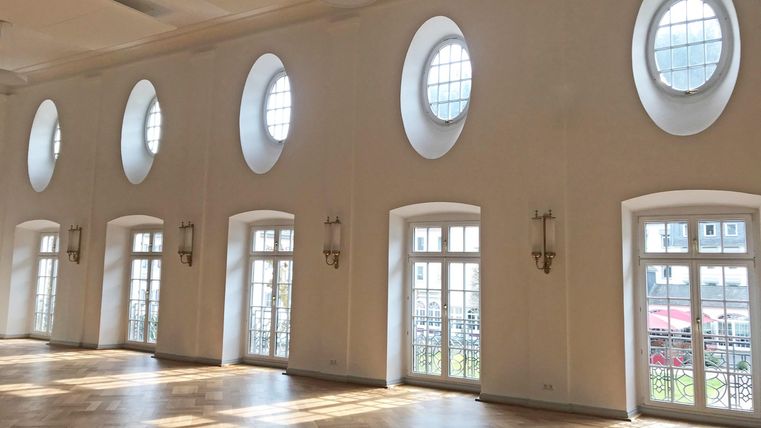 Een lichte kamer met ovale ramen en grote deuren. Daglicht valt op de elegante houten vloer.