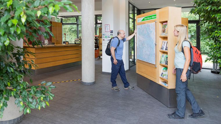 Ein Empfangsbereich mit zwei Personen, die an einer Informationswand stehen. Im Hintergrund sind Pflanzen und eine Holztheke zu sehen.