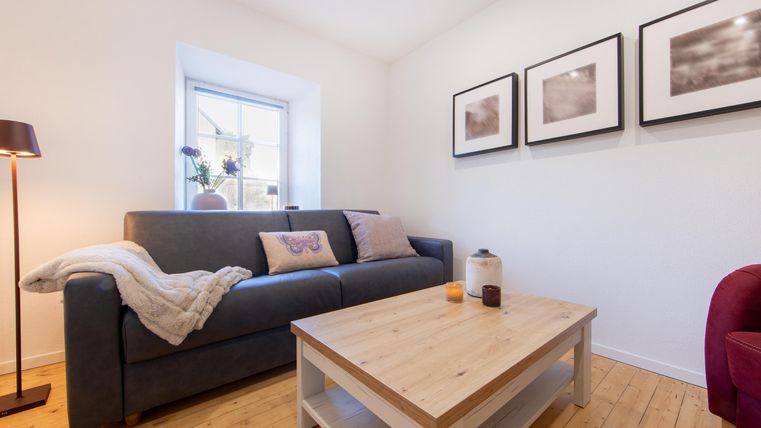 Ein gemütliches Wohnzimmer mit einem grauen Sofa und einem Holztisch. An der Wand hängen mehrere Bilder, und es gibt eine Lampe in der Ecke.