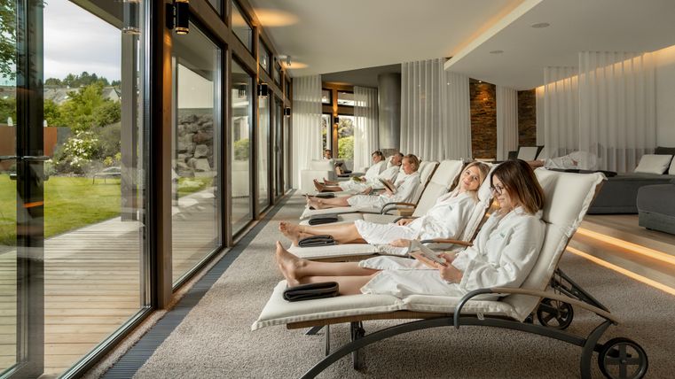 Ein entspannender Wellnessbereich mit bequemen Liegen. Fünf Personen in Bademänteln genießen die Ruhe und Aussicht.