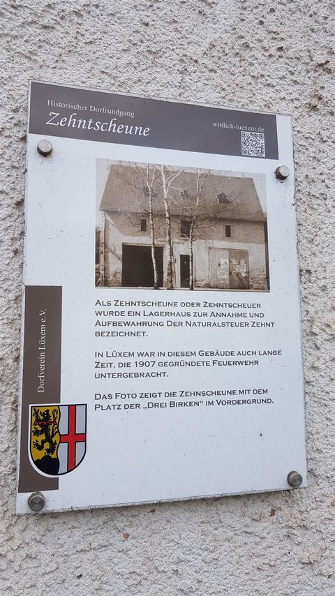 Ein Informationsschild an einer Wand zeigt ein historisches Gebäude. Es enthält Details zur Geschichte des Ortes und ist mit einem Wappen versehen.