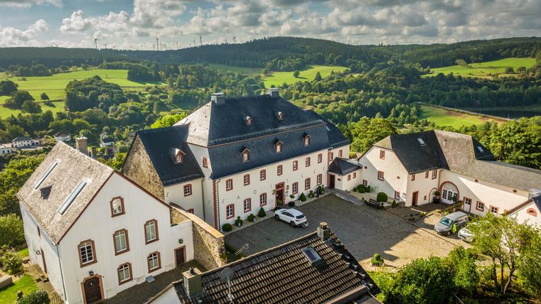 Een schilderachtig landhuis omringd door groene heuvels en bomen. De lucht is helder met enkele wolken.