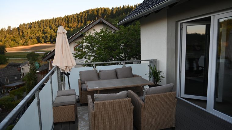 Een gezellige terras met een eettafel en stoelen van rattan. Op de achtergrond zijn bomen en heuvels te zien die een rustige landschap vormen.