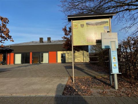 A modern building with an information board in the foreground. The surroundings are autumnal with few leaves on the trees.