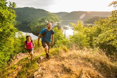 Zwei Wanderer auf einem Pfad mit Blick auf einen Stausee und bewaldete Hügel im Hintergrund.