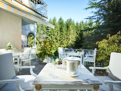 Een elegante terras met witte meubels en uitzicht op groene bomen. Een tafel is gedekt met een zilveren koelcontainer en verse bloemen.