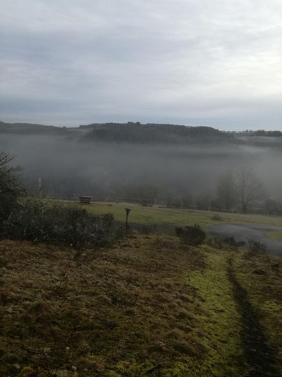Een mistig landschap met zachte heuvels op de achtergrond. De lucht is grijs en de sfeer lijkt rustig.