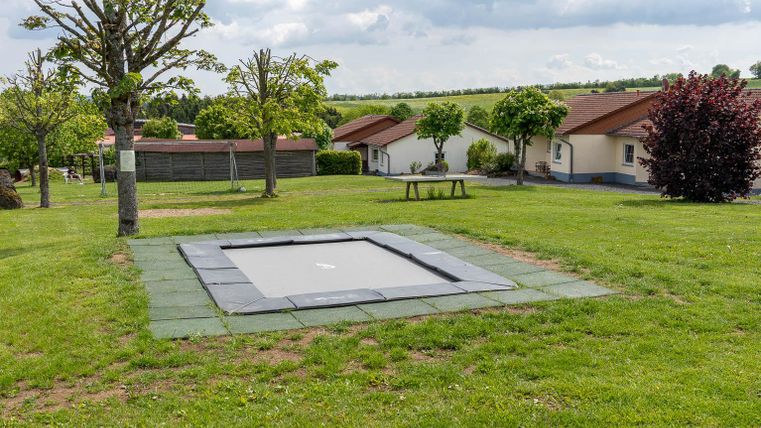 Een trampoline op een groene weide, omringd door bomen en huizen. De lucht is bewolkt, en de omgeving lijkt rustig en uitnodigend.