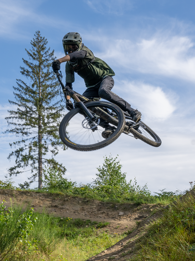 Een mountainbiker springt met zijn fiets over een heuvel. Op de achtergrond zijn bomen en een blauwe lucht te zien.