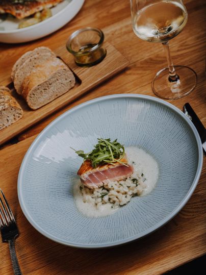Ein Teller mit gebratenem Fisch auf cremigem Risotto, garniert mit frischem Rucola. Dazu ein Glas Weißwein und frisches Brot auf dem Tisch.