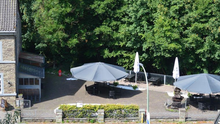 A cozy terrace with large umbrellas and seating. In the background, green trees can be seen.