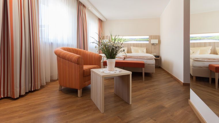 Ein modernes Zimmer mit Holzfußboden, einem orangefarbenen Sessel und einem Tisch mit einer Pflanze. Die großen Fenster sorgen für viel Tageslicht und eine freundliche Atmosphäre.