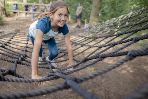 Een meisje klimt in een net in het bos. Ze glimlacht en lijkt veel plezier te hebben.