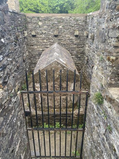 Steinernes Denkmal hinter einem Metallgitter, umgeben von alten Steinmauern und grüner Vegetation im Hintergrund.