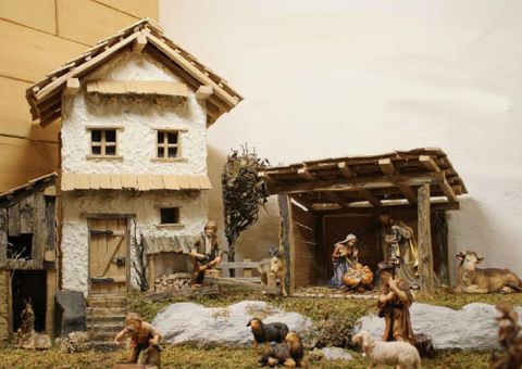 Eine traditionelle Krippe mit einem hübschen, detailreichen Stall und einem kleinen Haus. Figuren von Hirten, Tieren und der Heiligen Familie sind im Vordergrund zu sehen.