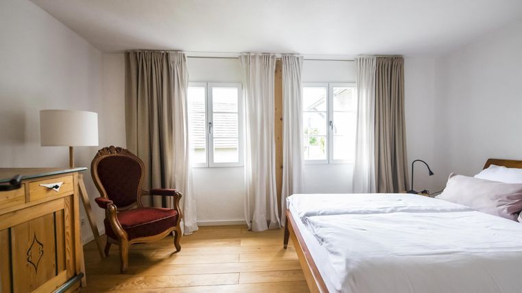 Ein helles Schlafzimmer mit zwei Betten und großen Fenstern. Ein bequemer Sessel und ein Holzschrank ergänzen die gemütliche Einrichtung.