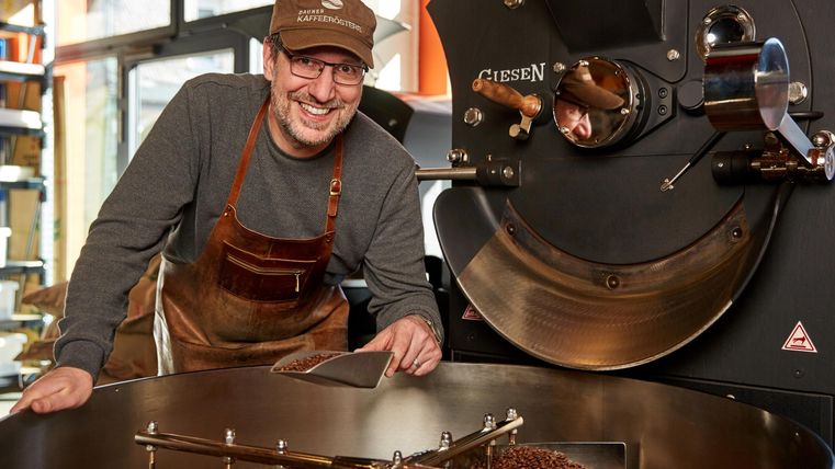 Ein Kaffeeröster lächelt neben einer großen Röstmaschine. Kaffeebohnen sind in der Maschine sichtbar.