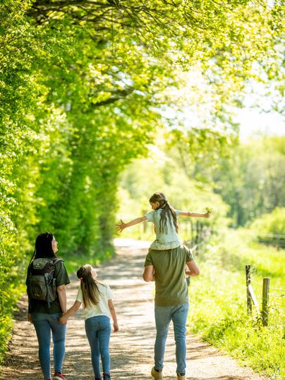 Een gezin loopt langs een zonnig pad, omgeven door groene bomen. Een kind zit op de schouders van een ouder en de anderen houden hand in hand.