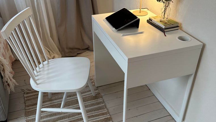 Een eenvoudige bureau met een lamp, een tablet en een kleine vaas met bloemen. Naast het bureau staat een houten stoel en op de vloer ligt een tapijt.