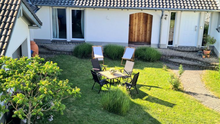 Ein gemütlicher Garten mit einem Tisch und Stühlen. Im Hintergrund steht ein Haus mit einer Holztür und großen Fenstern.