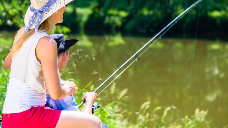Zwei sommerliche gekleidete Personen sitzen nebeneinander am Ufer eines Gewässers und halten Angeln in das Wasser. Um sie herum Wiese und Wälder.