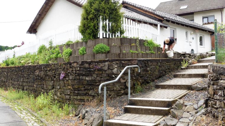 Ein kleines Haus mit einem Garten ist von einer Steinmauer umgeben. Eine Person gießt Pflanzen auf der Treppe.