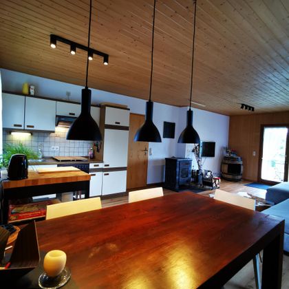 Een moderne keuken met een grote houten eettafel en zwarte verlichting. De ruimte is licht en vriendelijk, met toegang tot een buitenruimte.