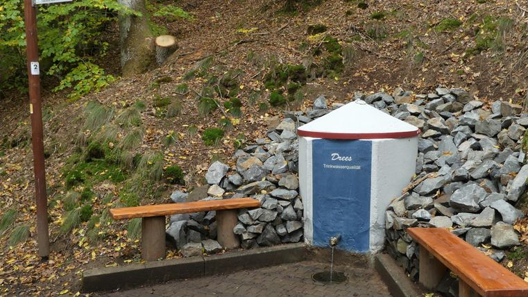 A small water fountain in nature, surrounded by stones and benches. The surroundings are green and show a lot of earth and plant soil.