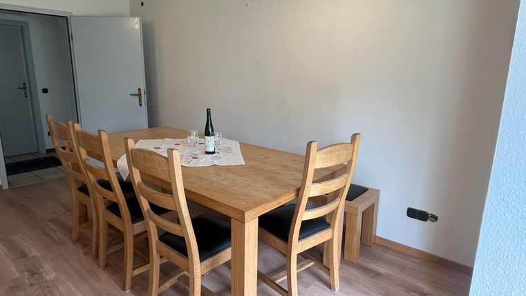 A bright dining room with a large wooden table and six chairs. On the table, there is a bottle of wine and a table runner.
