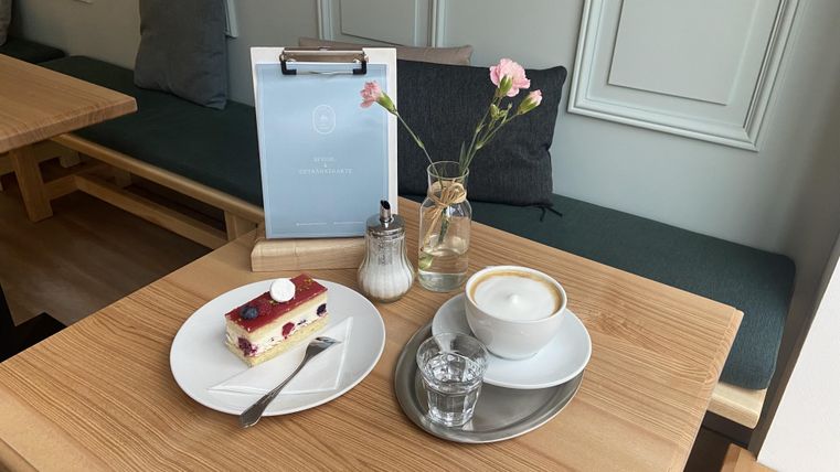 Een tafel in een café met een stuk taart en een kop koffie. Een kleine vaas met een bloem staat naast het dessert.