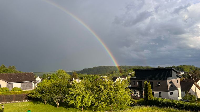 A rainbow appears over a tranquil landscape with houses and green trees. The sky is cloudy, but the colors of the rainbow stand out clearly.
