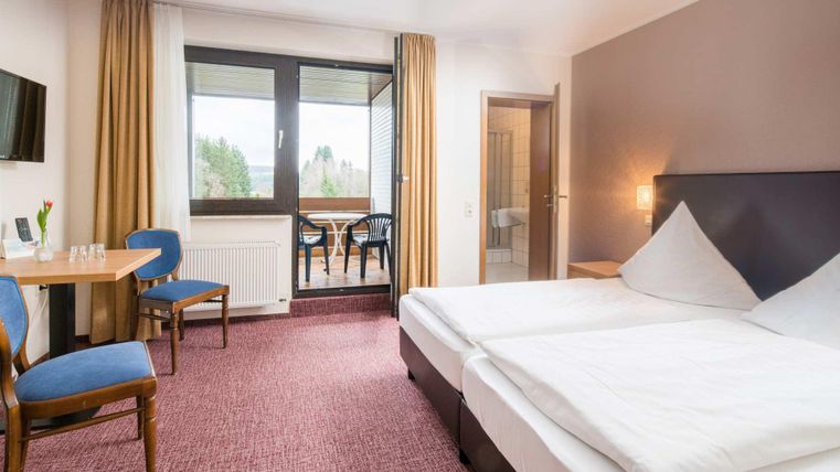 A modern hotel room with two single beds, a table, and chairs. Large windows offer a view of the outdoor area.