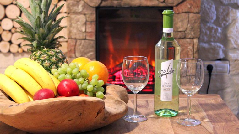 Ein Holztablett voller frischer Früchte wie Bananen, Äpfel und Trauben steht auf einem Tisch. Neben dem Tablett befindet sich eine Flasche Weißwein und zwei Weingläser vor einem gemütlichen Kamin.