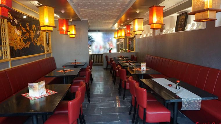 Ein modernes Restaurant mit roten Sitzbänken und dunklen Tischen. Die Wände sind mit asiatischen Dekorationen und leuchtenden Laternen geschmückt.