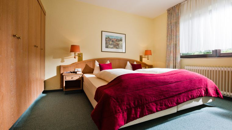 Een moderne slaapkamer met een groot bed en rode dekbedden. De muren zijn geschilderd in een zachte gele kleur en er is een kast en een nachtkastje.