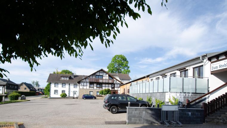 Ein ruhiger Parkplatz vor einem Hotel mit mehreren Gebäuden. Der Himmel ist klar und sonnig, umgeben von Bäumen.