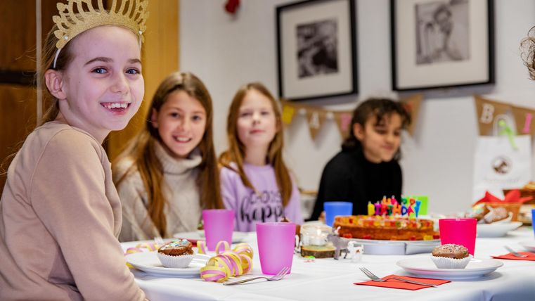 Eine fröhliche Gruppe von Mädchen feiert zusammen an einer geschmückten Tafel. Auf dem Tisch steht ein Geburtstagskuchen mit Kerzen und bunte Getränke.