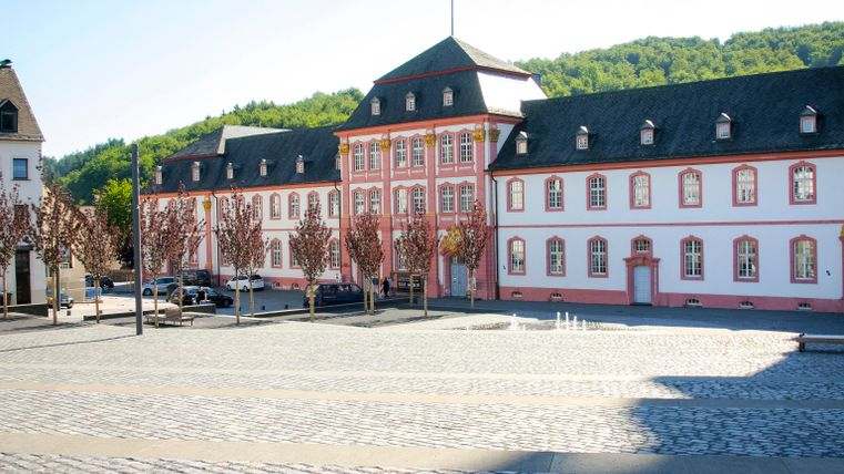 A historic building with red and white masonry stands in a square. In the background, green hills and a clear sky can be seen.