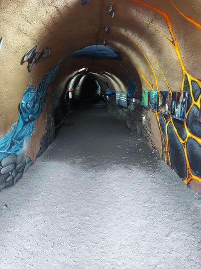 Een lange, smalle tunnel met kleurrijke muurschilderingen. De vloer is grotendeels steenslag en er zijn kunstwerken aan de muren.