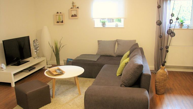 A cozy living room with a gray sofa and a round table. The room has a television and bright windows with plants.