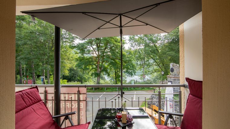 Ein gemütlicher Balkon mit zwei roten Stühlen und einem Tisch. Im Hintergrund sind Bäume und ein Blick auf das Wasser zu sehen.