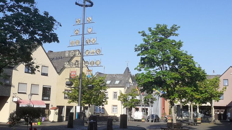 The Graf-Mirbach-Platz in Hillesheim with some shops and residential buildings around it and a guild tree in the middle.