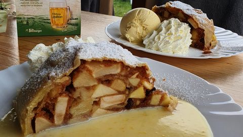 Ein Stück Apfelstrudel mit Zuckerguss und Vanillesoße steht auf einem Teller. Im Hintergrund sieht man eine Portion Eiscreme und Schlagsahne.
