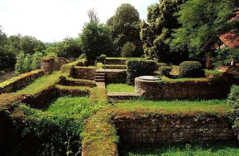 Ruinen einer alten Villa mit überwucherten Mauern und grüner Vegetation, umgeben von Bäumen und einem klaren Himmel.