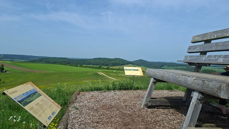 A wooden bench stands at a viewpoint with green fields and a blue sky in the background. Next to the bench, information boards are set up.