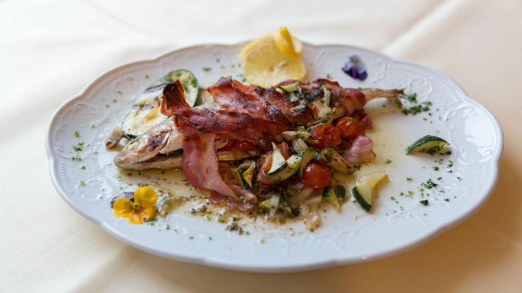 Ein liebevoll angerichteter Teller mit gegrilltem Fisch und knusprigem Speck. Das Gericht ist mit frischen Kräutern und Gemüse dekoriert.