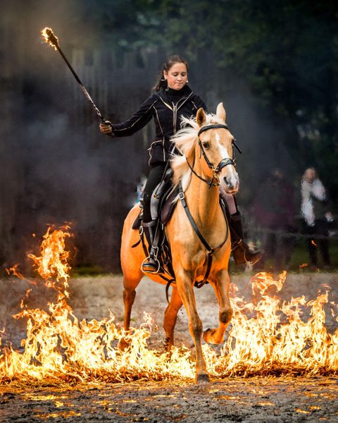 Een rijdster op een paard steekt een brandend gebied over. Ze houdt een brandende stok in haar hand.