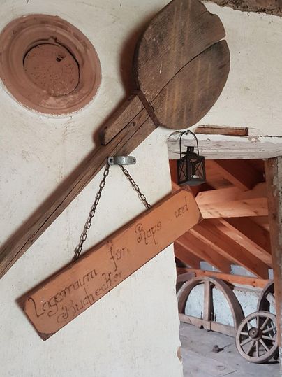 Ein rustikales Gebäude mit einem großen Holzlöffel und einer hängenden Lampe an der Wand. Ein Holzschild mit eingraviertem Text ist ebenfalls sichtbar.