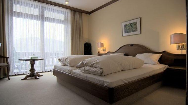 Een gezellige slaapkamer met een groot bed en lichte gordijnen. Een kleine tafel staat naast het bed en de kamer heeft een warme, uitnodigende uitstraling.