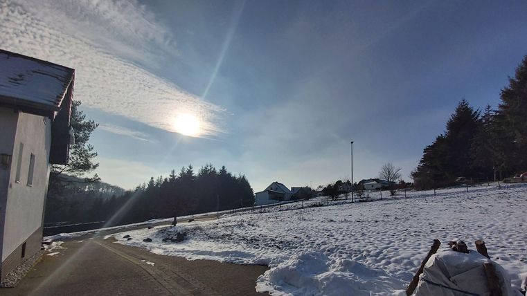 Een met sneeuw bedekt landschap onder een heldere lucht. De zon straalt over de bomen en weilanden.